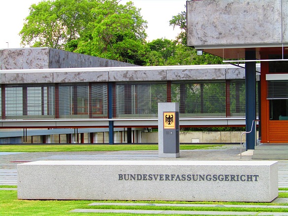 Ausschnitt des Gebäudes des Bundesverfassungsgerichts, davor ist grüner Rasen, vorne der Schriftzug und in der Mitte des Bildes der Bundesadler aus einem Schild. Das Gebäude besteht vor allem aus Beton und Glas.